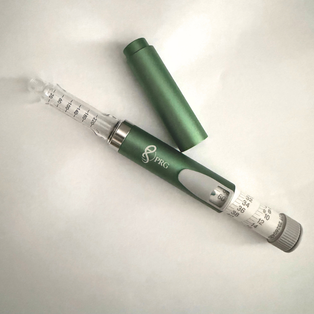 A green and silver PRG insulin pen with its cap removed, lying diagonally on a light off-white background.